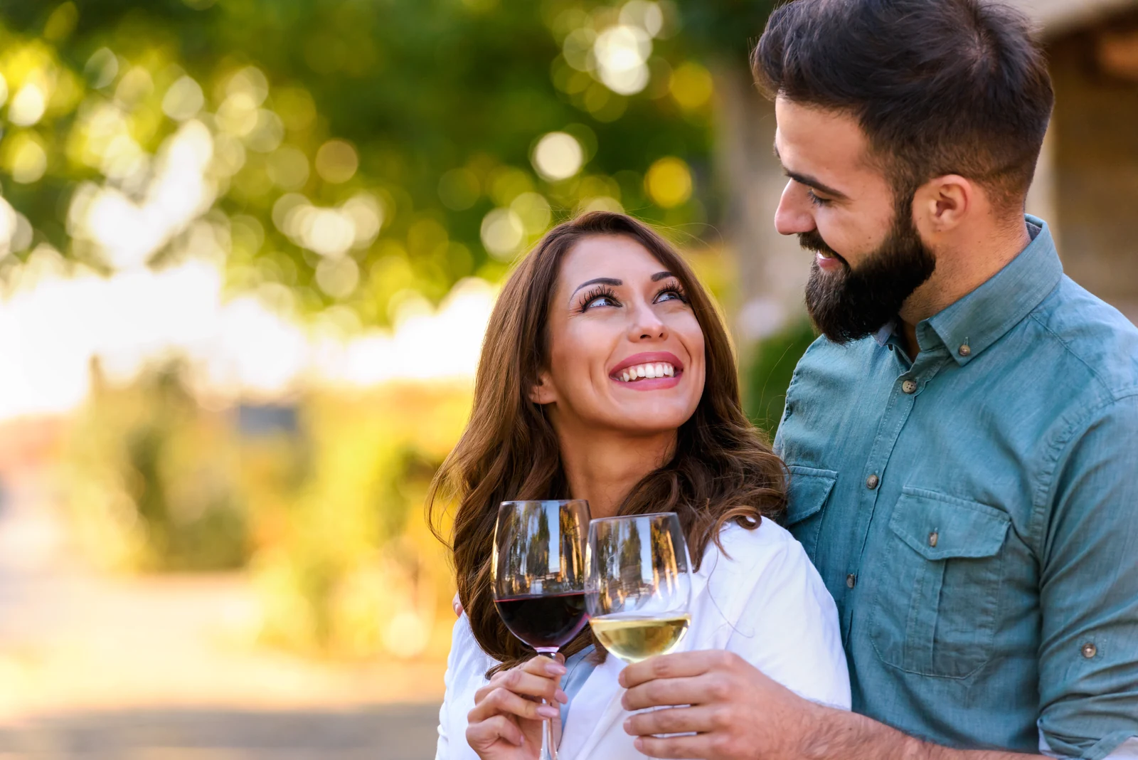 Smiling couple toasting with red and white wine in a sunny vineyard