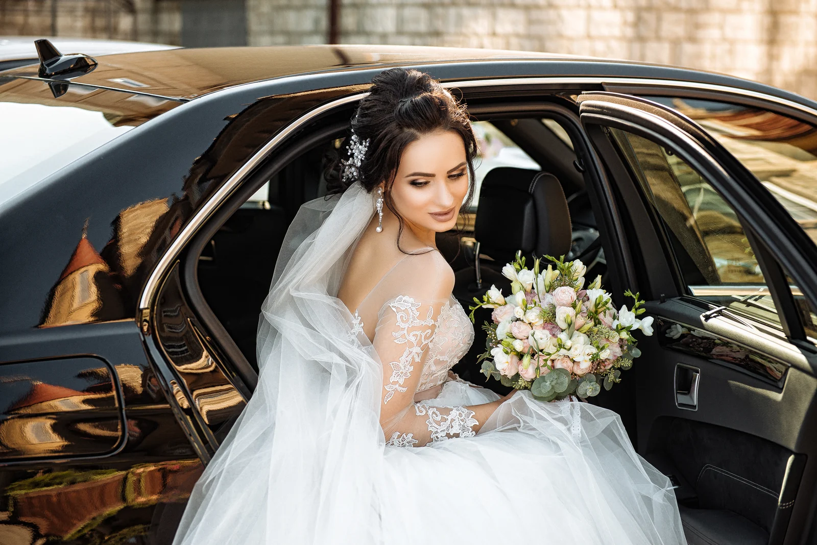 Smiling bride holding a bouquet in the open rear door of a black wedding car