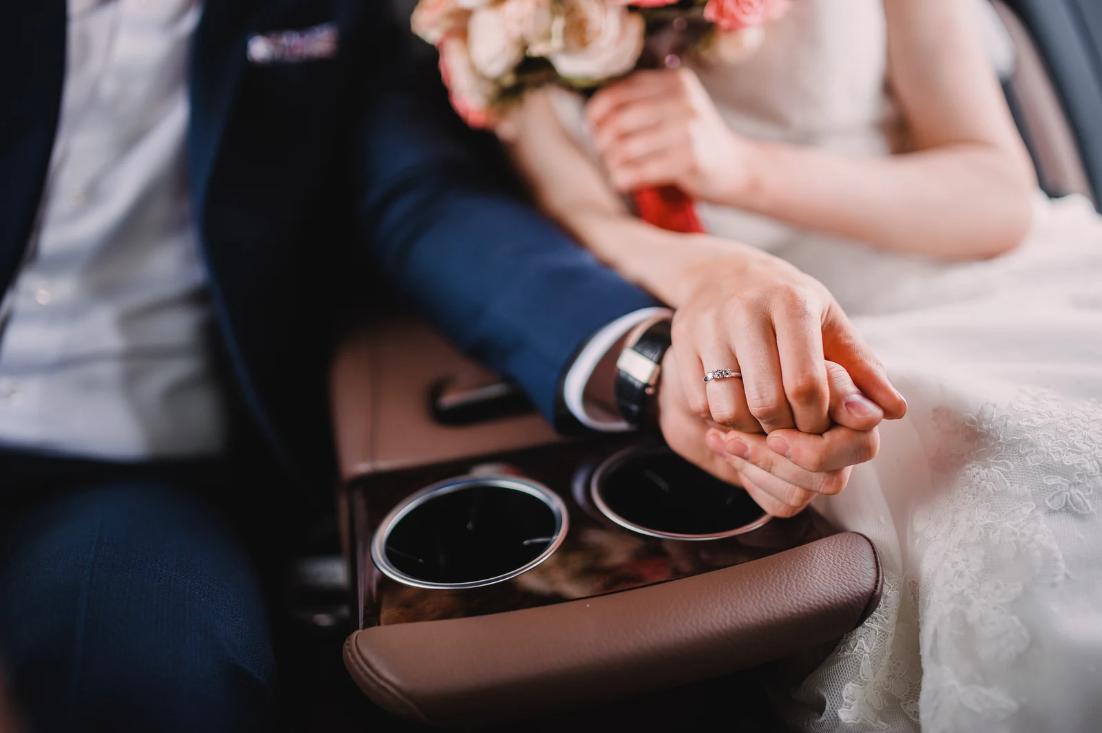Bride and groom holding hands in the back seat of a wedding car, wedding ring in focus