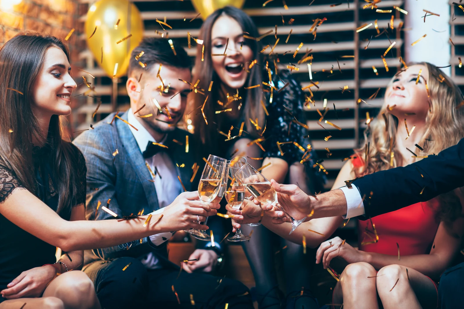 Friends in evening wear raising champagne glasses as gold confetti falls at a celebration