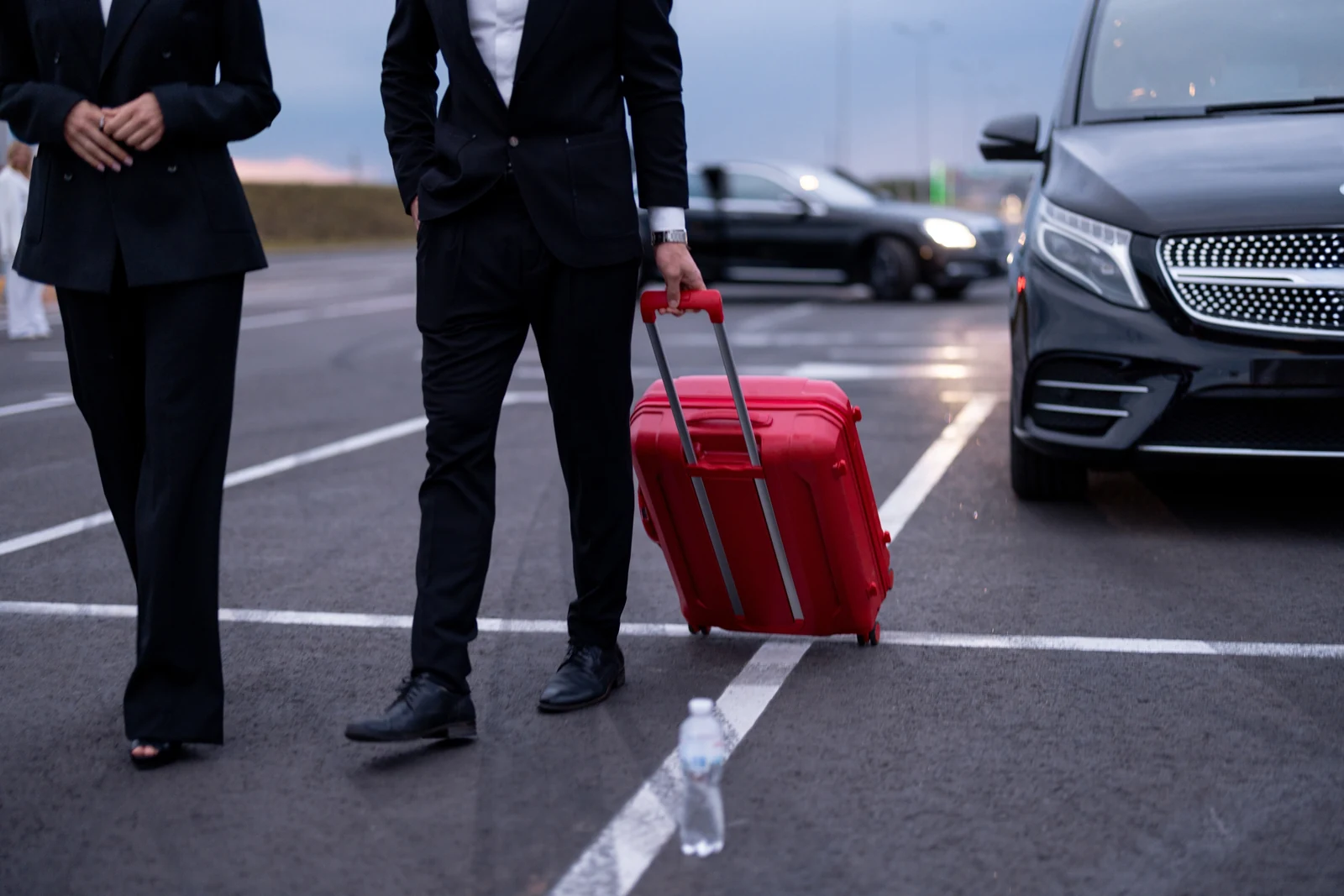 Two business travellers in dark suits walking with a red suitcase towards a black Mercedes-Benz V-Class
