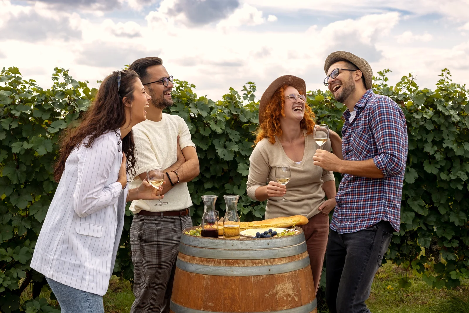 Two couples laughing with wine around a barrel in a vineyard