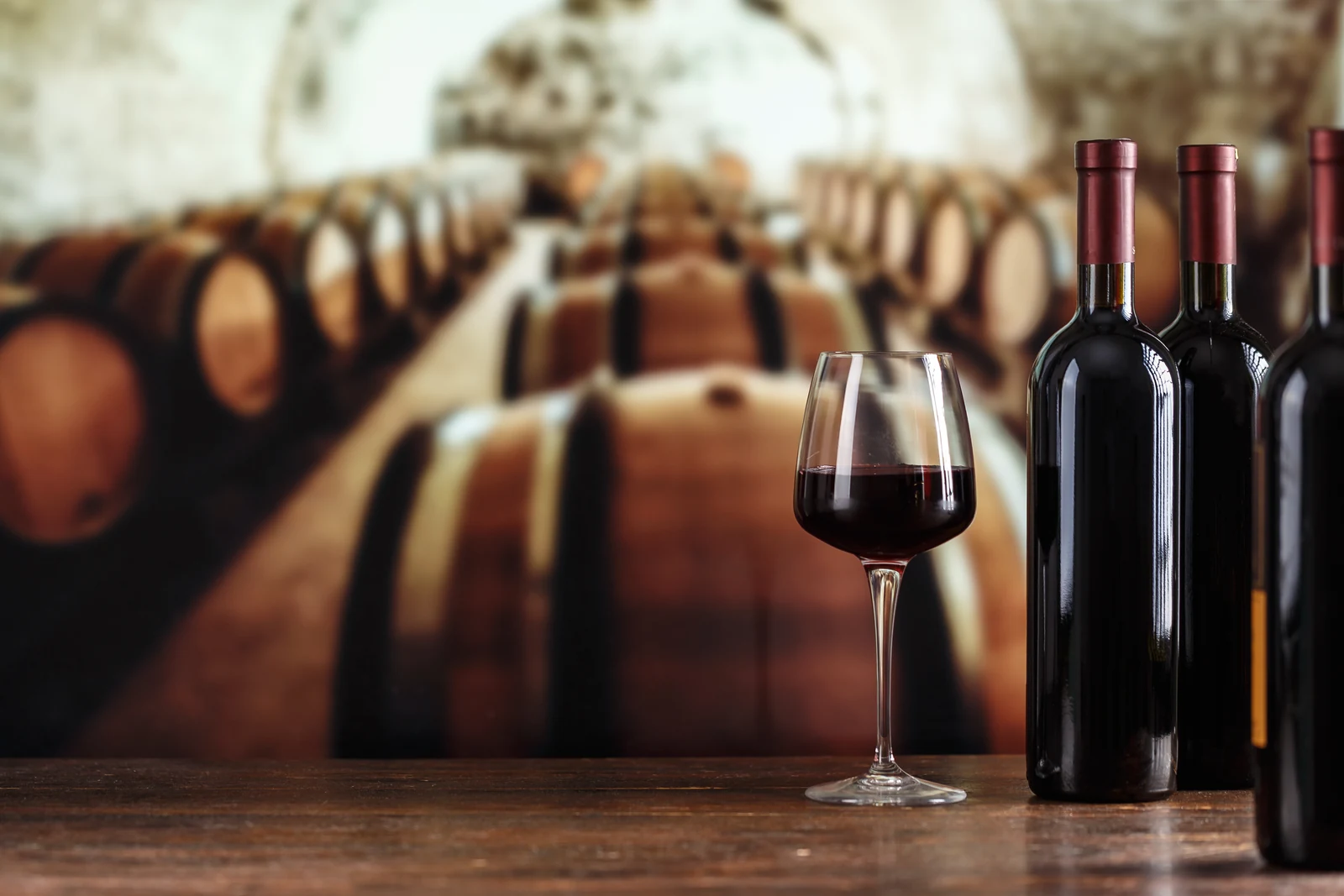 Glass of red wine and bottles on a table in front of an oak barrel cellar